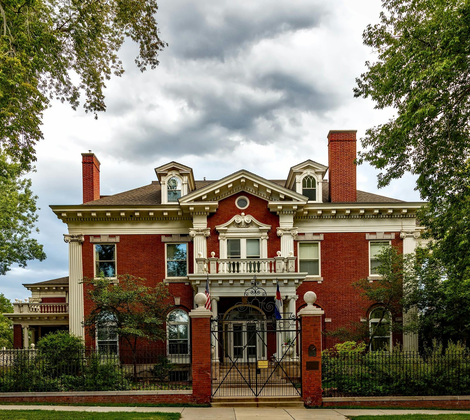Historic red brick mansion with a grand entrance and ornate gate under a cloudy sky.